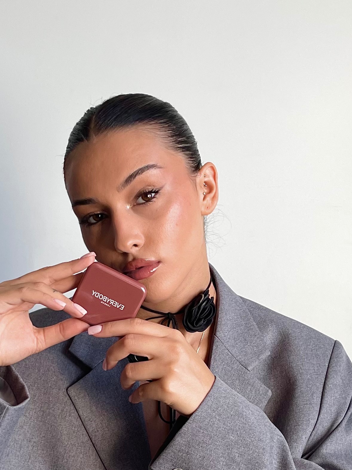 Portret kobiety w szarym żakiecie, prezentującej kosmetyk EverybodyLondon. Makijaż z błyszczącymi ustami i delikatnym oświetleniem podkreśla efekt nowej kolekcji.