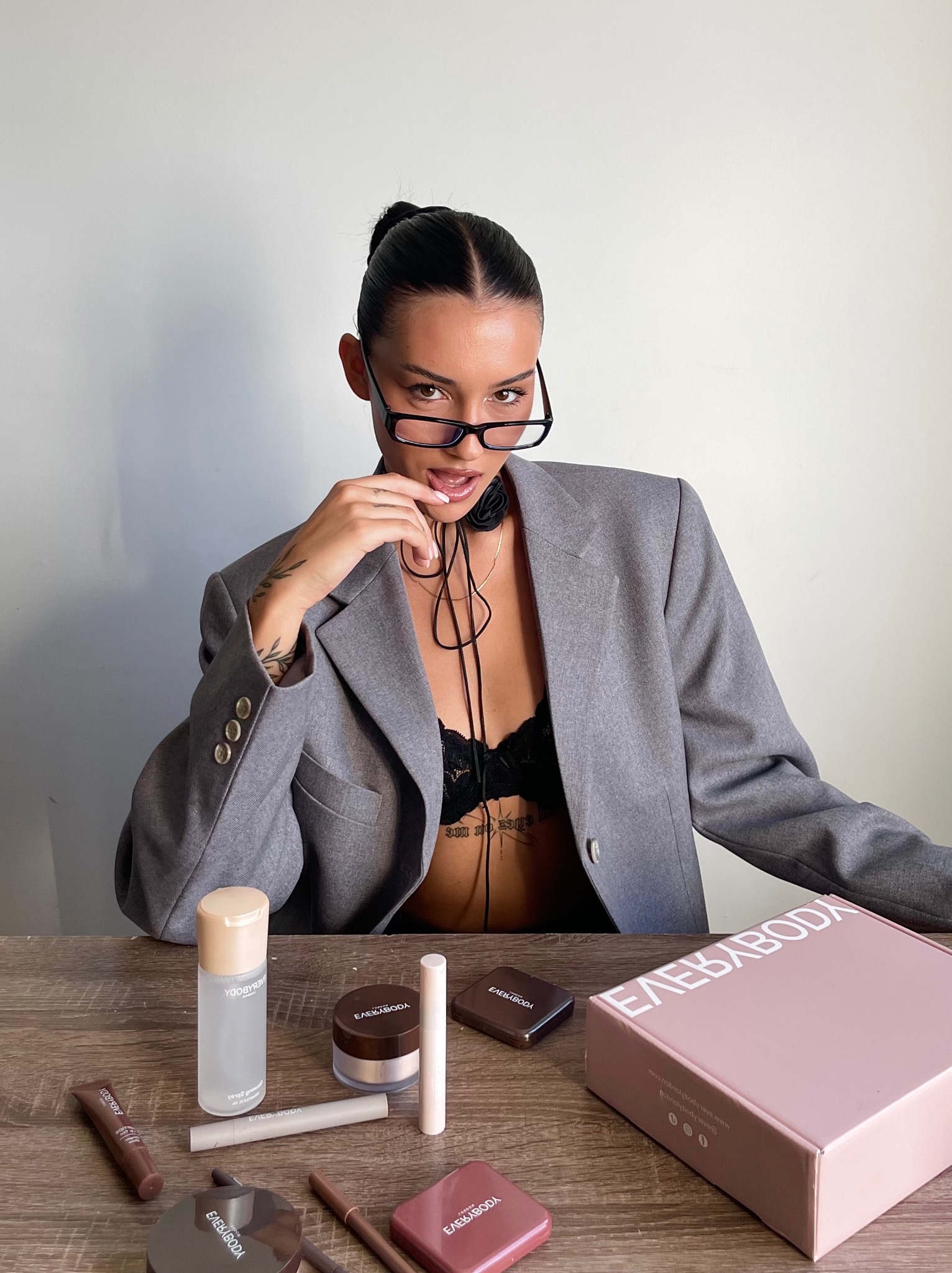 Sesja wizerunkowa marki kosmetycznej EverybodyLondon: modelka w okularach i marynarce prezentuje nową kolekcję kosmetyków na drewnianym stole.