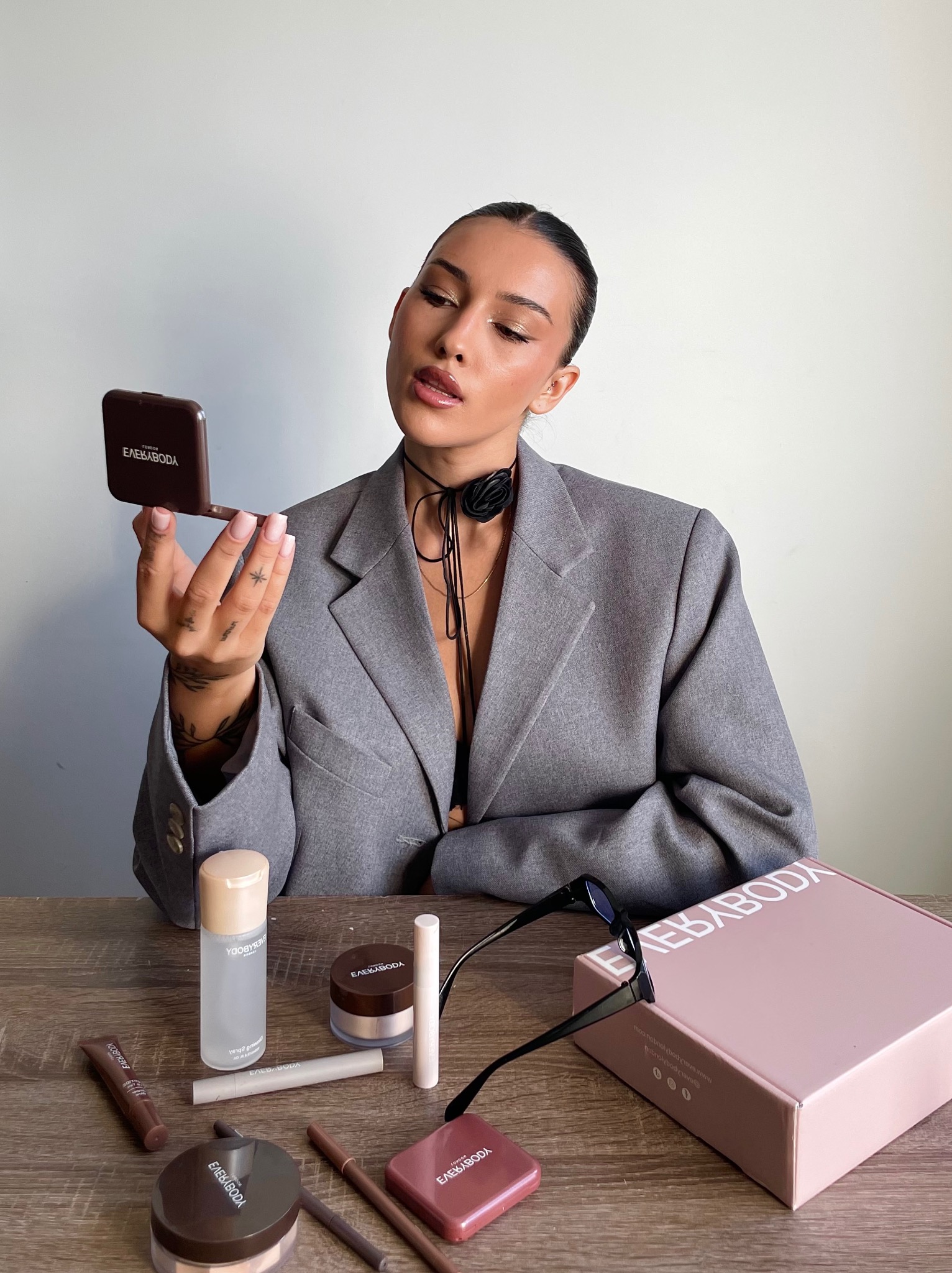 Sesja kosmetyczna EverybodyLondon: modelka w szarym żakiecie przegląda się w lusterku pudru, na drewnianym stole rozłożone kosmetyki i pudełko z logo.