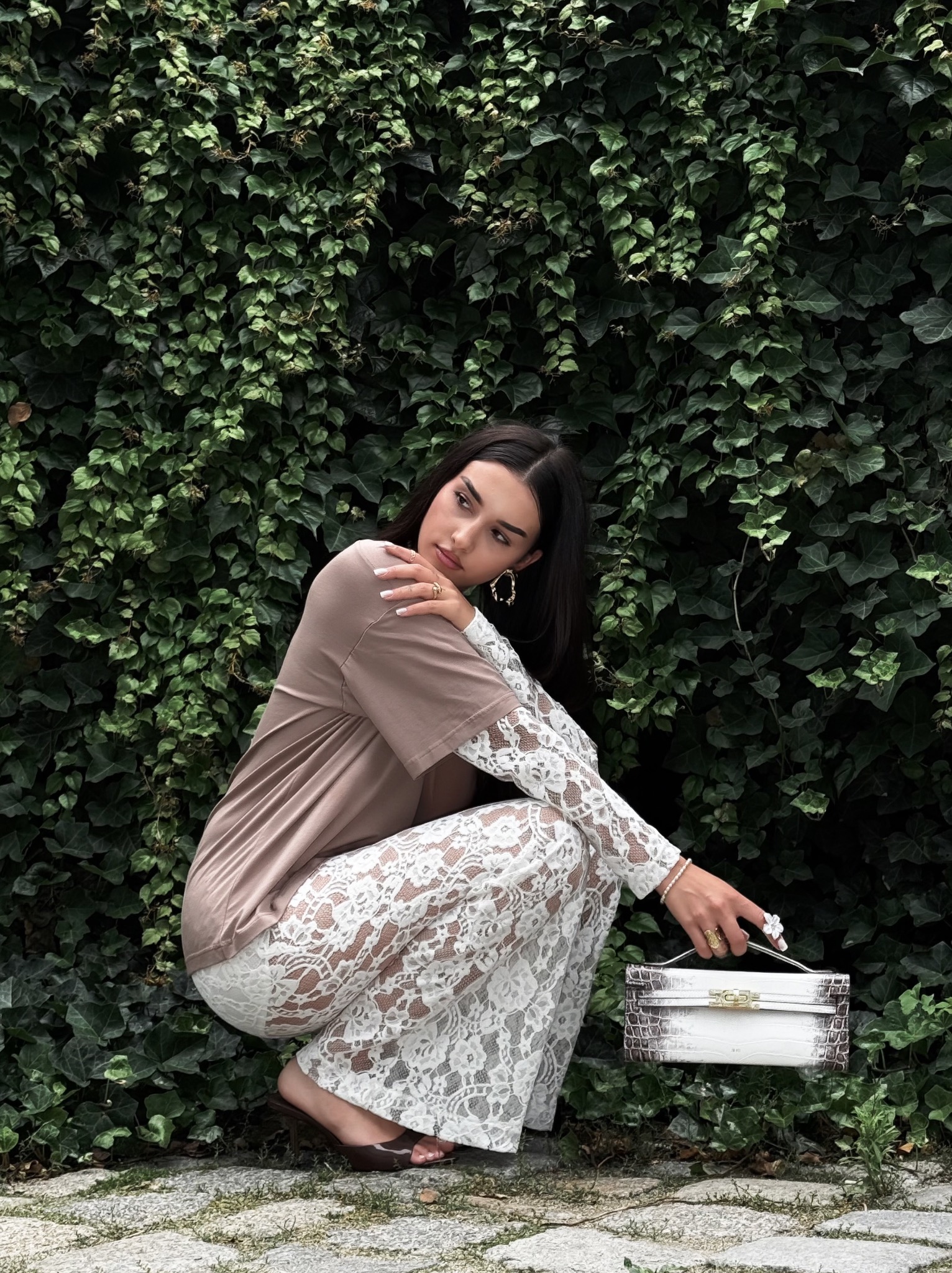 Modelka w koronkowej stylizacji prezentuje torebkę JWPEI na tle ściany z bluszczu. Ujęcie z dołu podkreśla detale stroju i akcesorium.