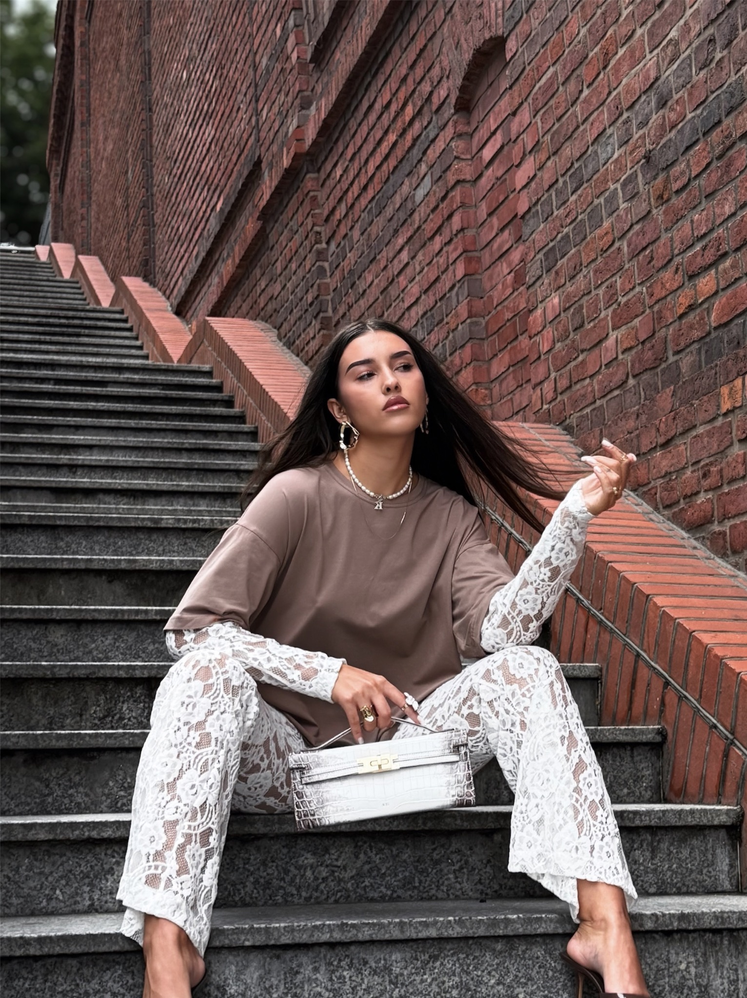 Modelka siedzi na schodach z torebką JWPEI, prezentując nową kolekcję. Ceglana ściana w tle, naturalne światło.