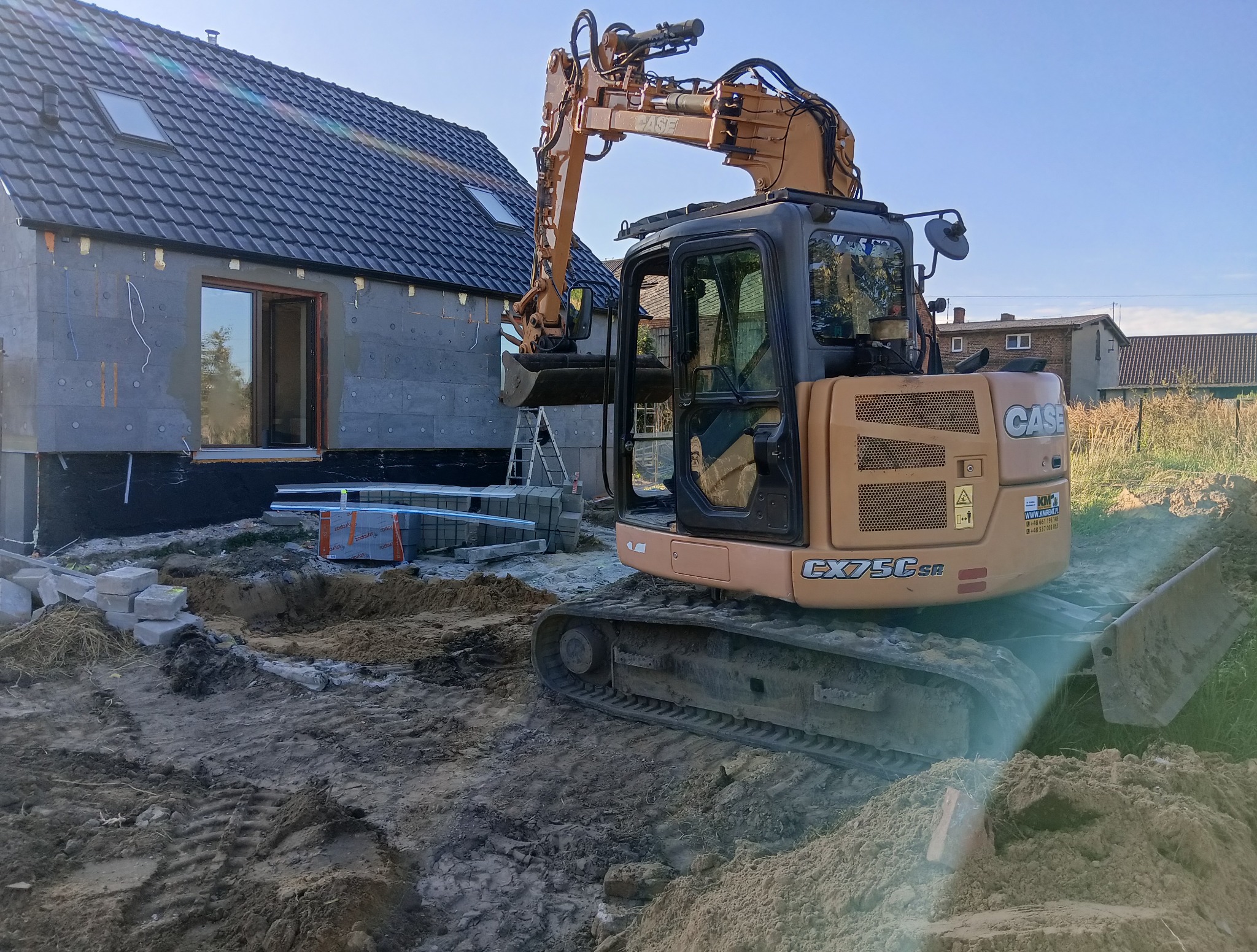 Żółta koparka CASE CX75CSR na placu budowy, częściowo wykopany fundament domu z szarym ociepleniem. Widoczne bloczki i materiały budowlane.