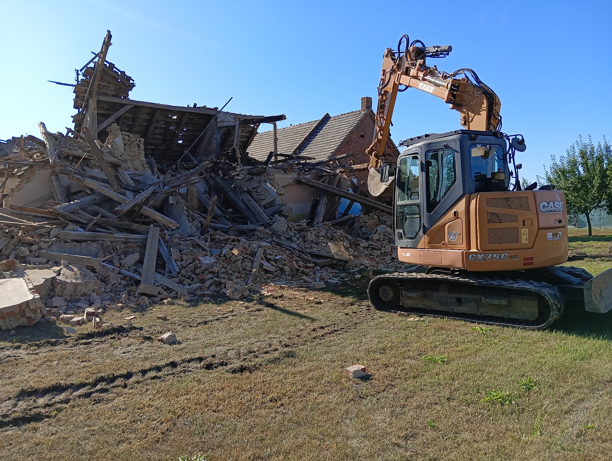 Pomarańczowa koparka CASE CX75C SR na tle sterty gruzu po rozbiórce budynku z cegły i drewna, widok z boku, słoneczny dzień.
