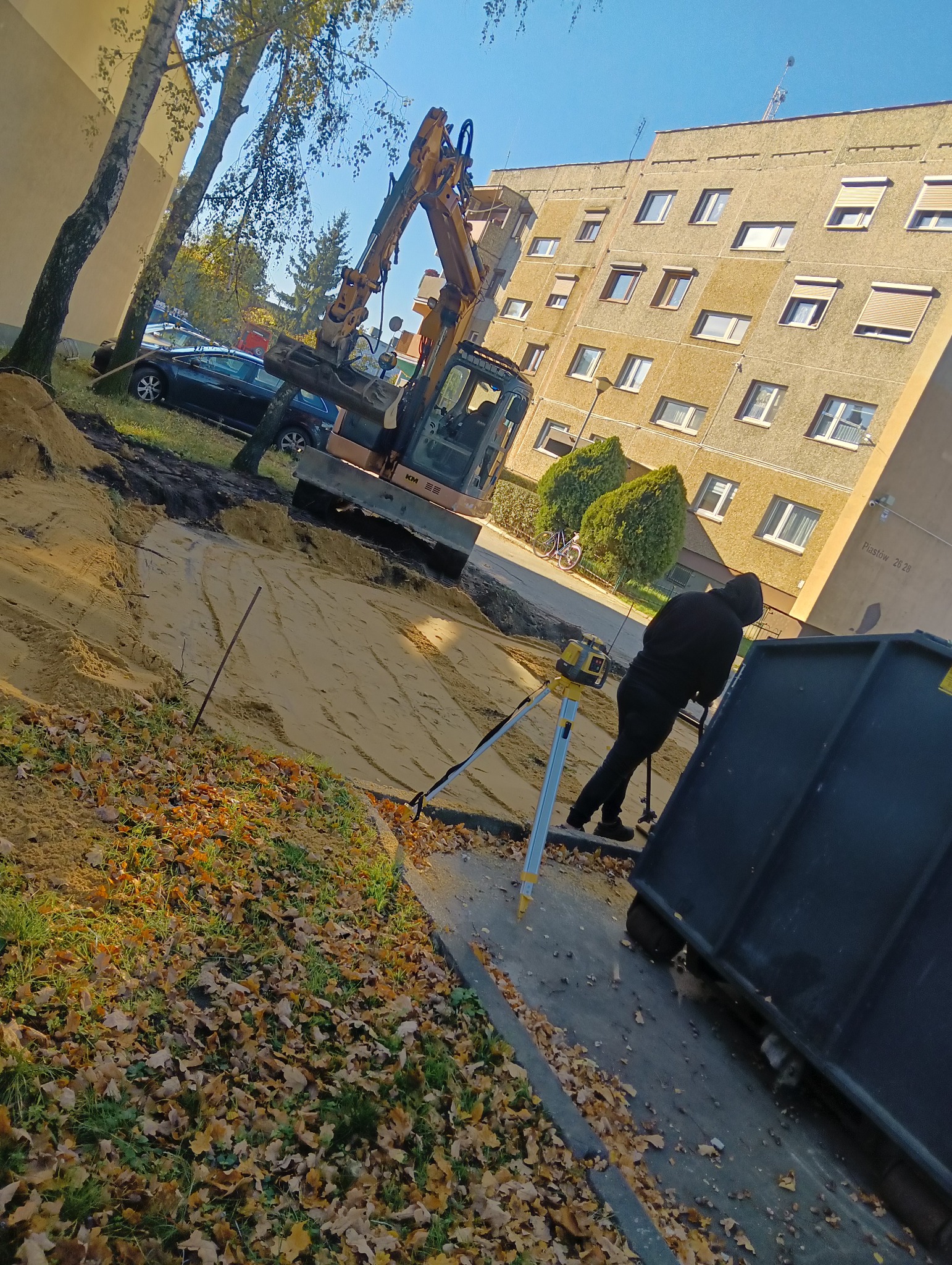 Żółta koparka KM na piasku, obok pracownik z niwelatorem. W tle blok mieszkalny i drzewa. Prace ziemne w toku, widoczny kontener na ziemię.