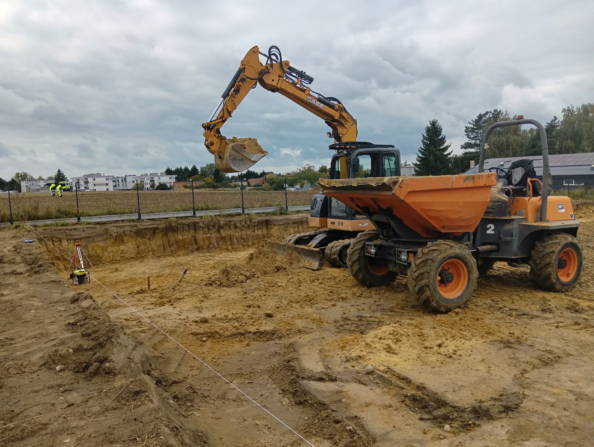 Żółta koparka nabiera ziemię do pomarańczowej wywrotki na wykopie pod fundamenty. W tle budynki mieszkalne i osoby w odblaskowych ubraniach.