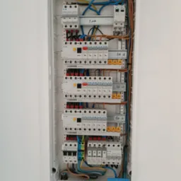 Rozdzielnica elektryczna w białej obudowie, widoczne bezpieczniki, przewody w kolorach: niebieskim, brązowym, żółto-zielonym, ułożone wewnątrz skrzynki.