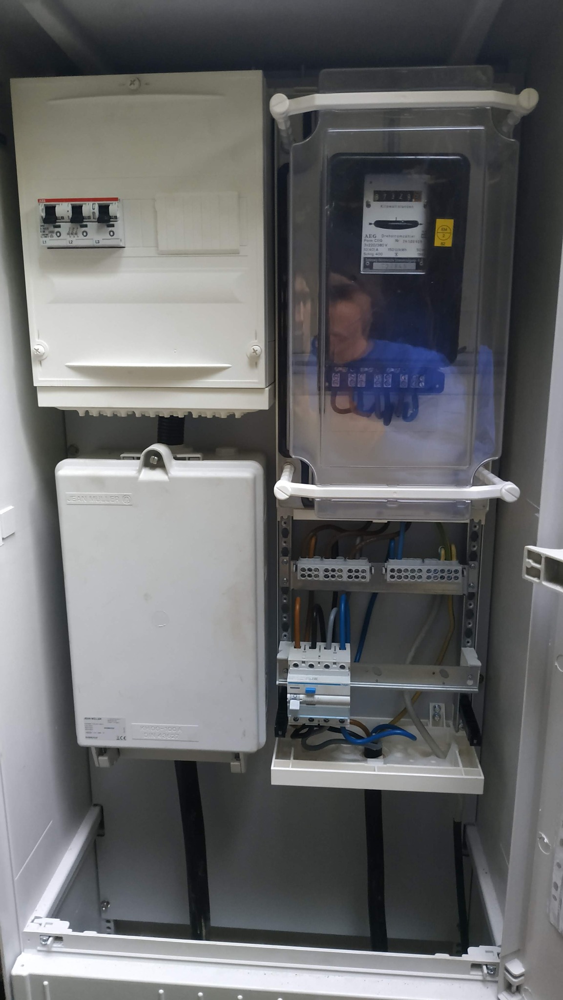 Rozdzielnica elektryczna z licznikiem i zabezpieczeniami, widoczne przewody i złączki. Skrzynka w kolorze białym, schludne ułożenie kabli.