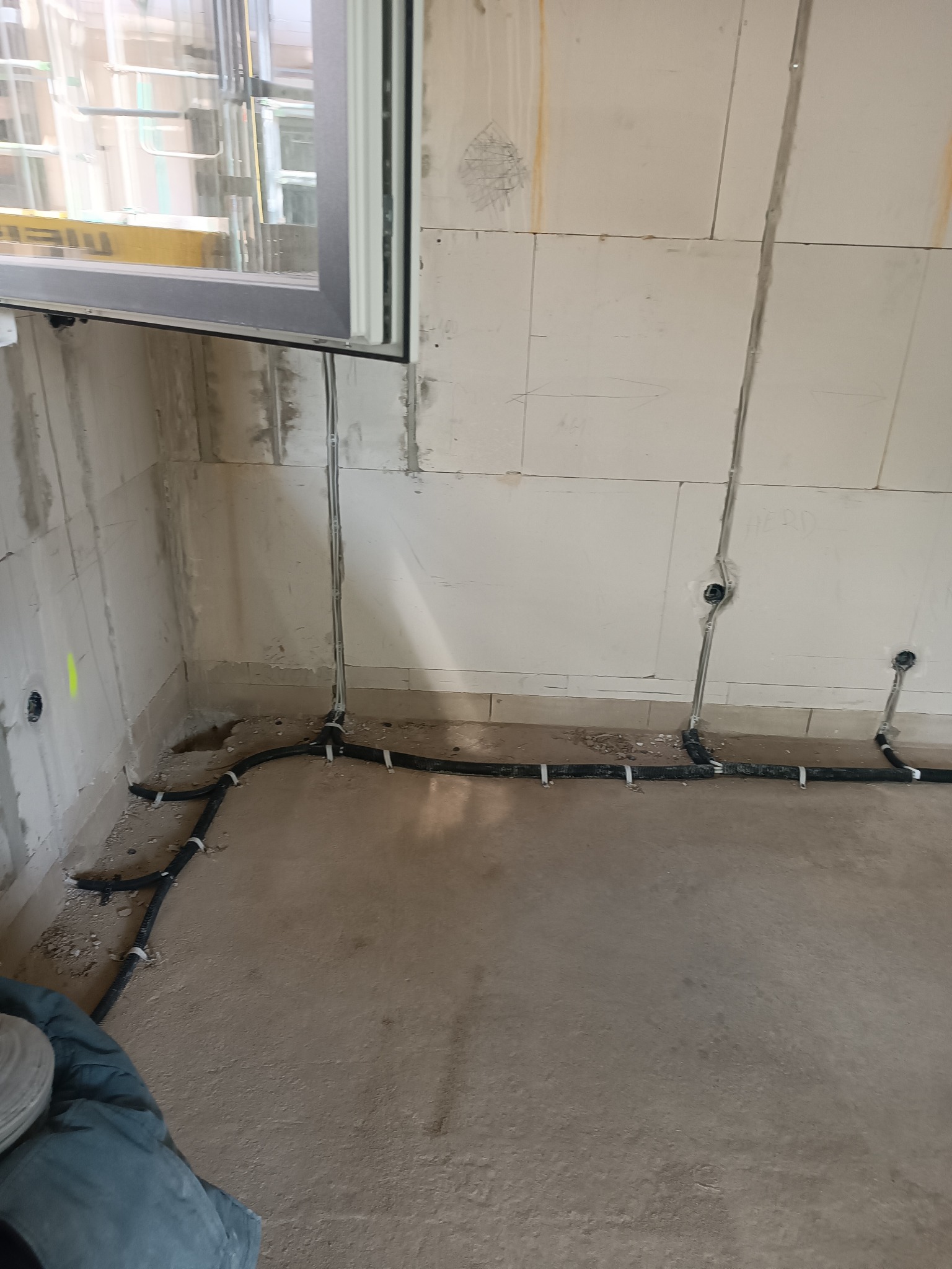 Wnętrze w trakcie prac wykończeniowych: widoczne przewody elektryczne poprowadzone wzdłuż ścian i podłogi, zamocowane uchwytami. Surowy stan ścian i okno w tle.