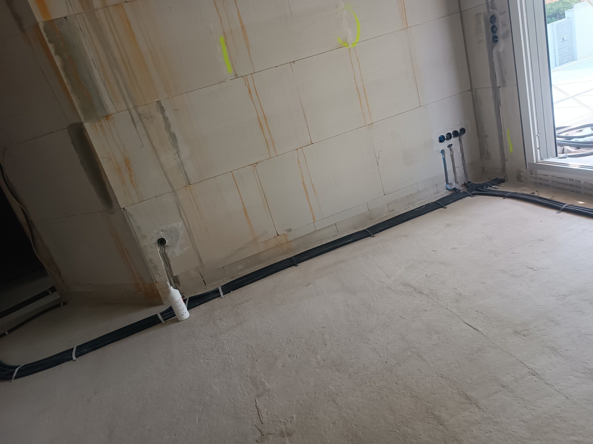 Surowa ściana z widocznymi przewodami elektrycznymi, rurkami i puszkami. Instalacja poprowadzona wzdłuż ściany, zabezpieczona opaskami. W tle okno z widokiem na zewnątrz.