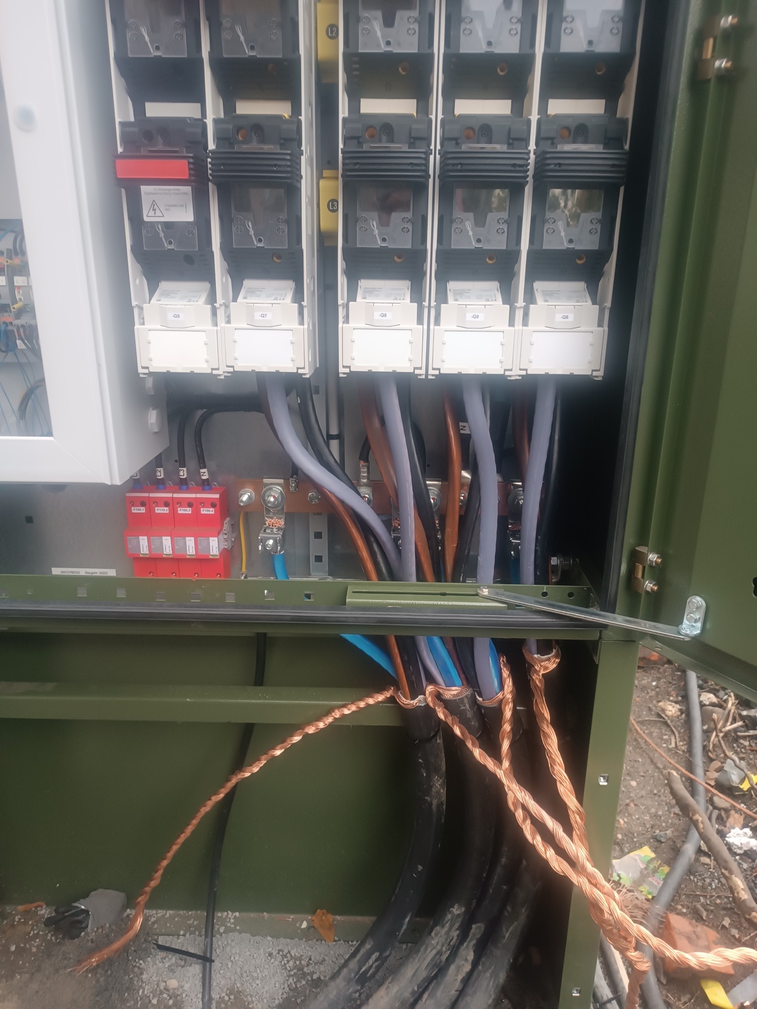 Otwarta skrzynka elektryczna w kolorze khaki z widocznymi bezpiecznikami, przewodami i elementami instalacji. Widoczne kable w oplocie miedzianym i plastikowe rury.