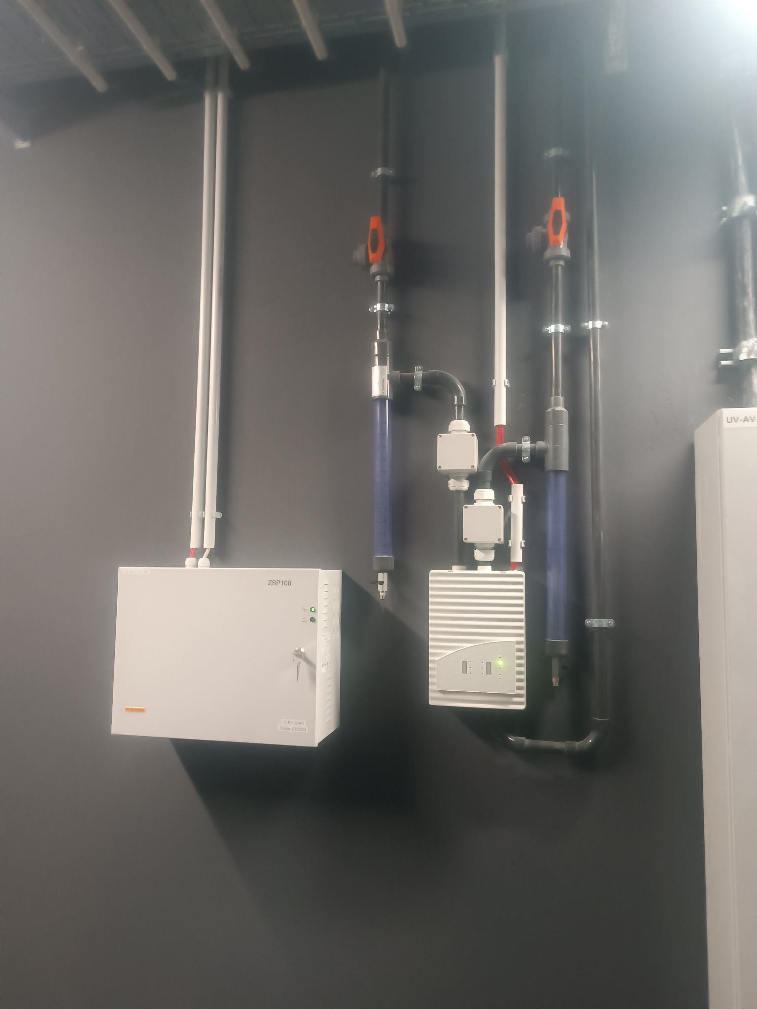 Szary korytarz z systemem uzdatniania wody: białe rury, zawory z pomarańczowymi uchwytami, skrzynki sterownicze. Widoczne elementy przyłączeniowe i wężyki.