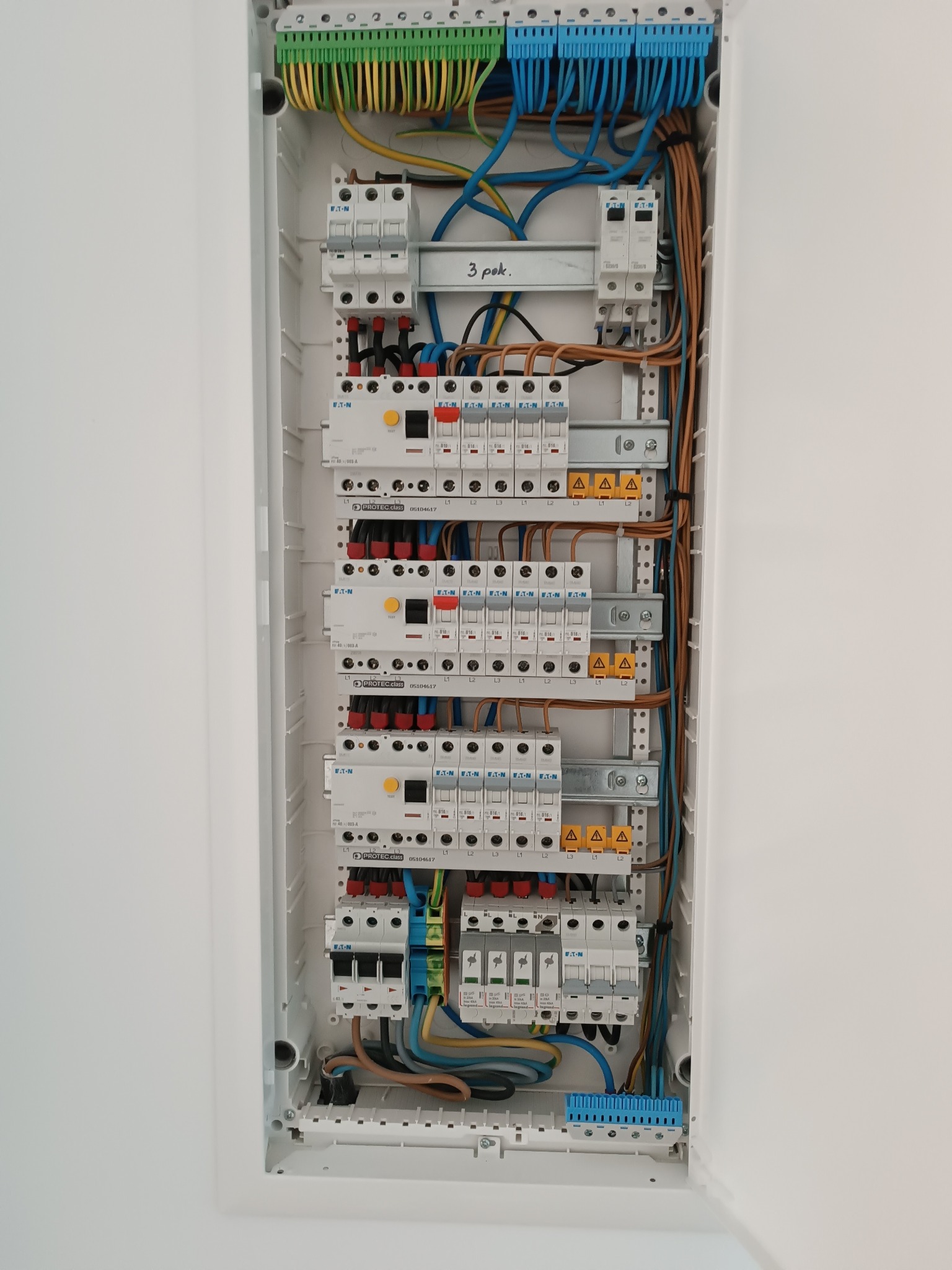 Rozdzielnica elektryczna w białej obudowie, widoczne bezpieczniki, przewody w kolorach: niebieskim, brązowym, żółto-zielonym, ułożone wewnątrz skrzynki.