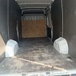 Pusta przestrzeń ładunkowa busa wyłożona płytami, widok z tyłu pojazdu, drzwi otwarte. Widoczna podłoga z zabrudzeniami i listwa przypodłogowa.