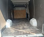 Pusta przestrzeń ładunkowa busa wyłożona płytami, widok z tyłu pojazdu, drzwi otwarte. Widoczna podłoga z zabrudzeniami i listwa przypodłogowa.