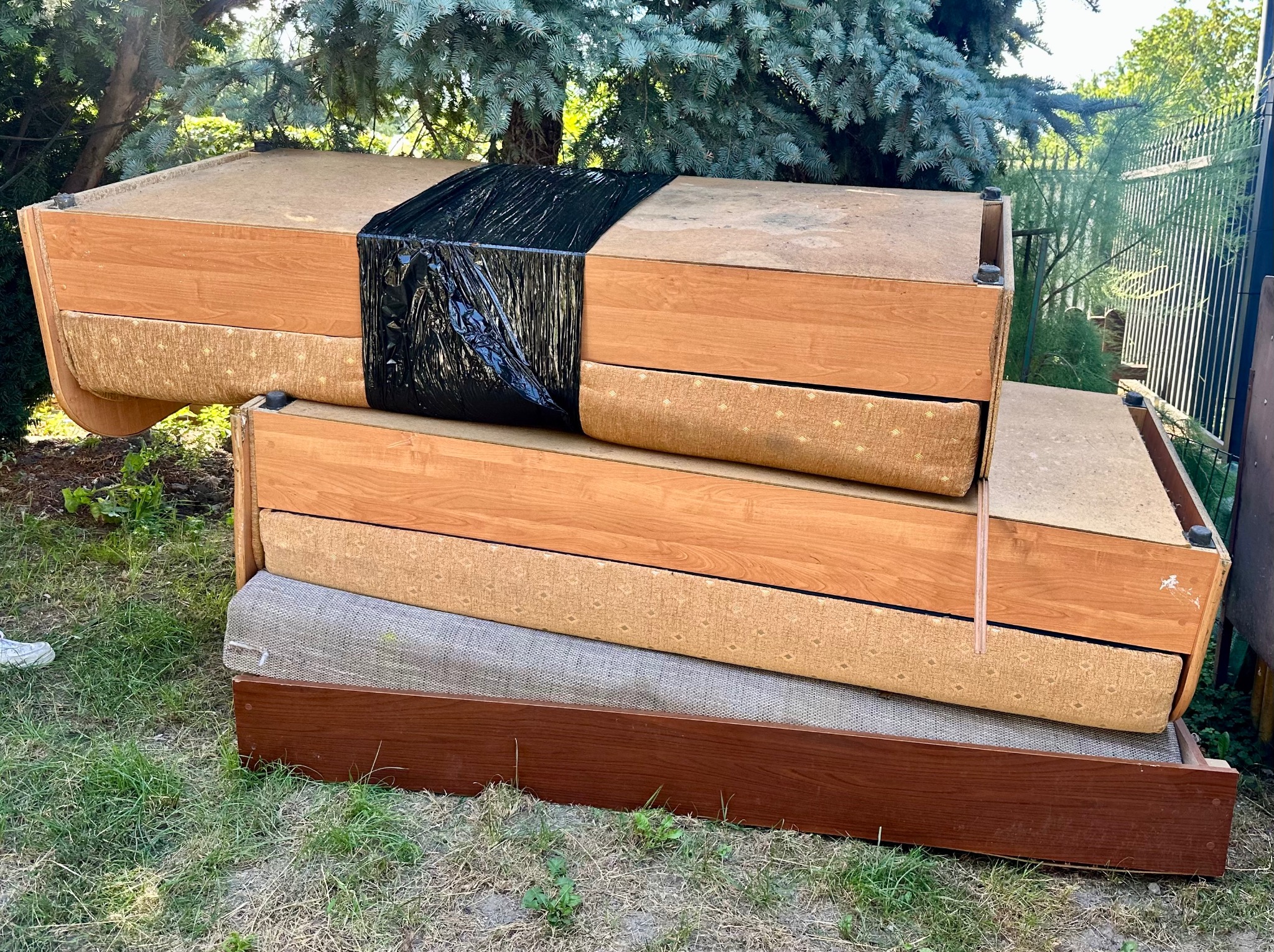 Stare, drewniane elementy meblowe, częściowo owinięte czarną folią, składowane na zewnątrz, na trawie, prawdopodobnie do wywiezienia podczas opróżniania mieszkania.