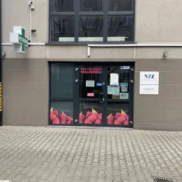 Wejście do przychodni Malinowa z plakatem malin na drzwiach, zielony krzyż apteczny i tabliczka NFZ na fasadzie budynku. Ujęcie z poziomu chodnika.
