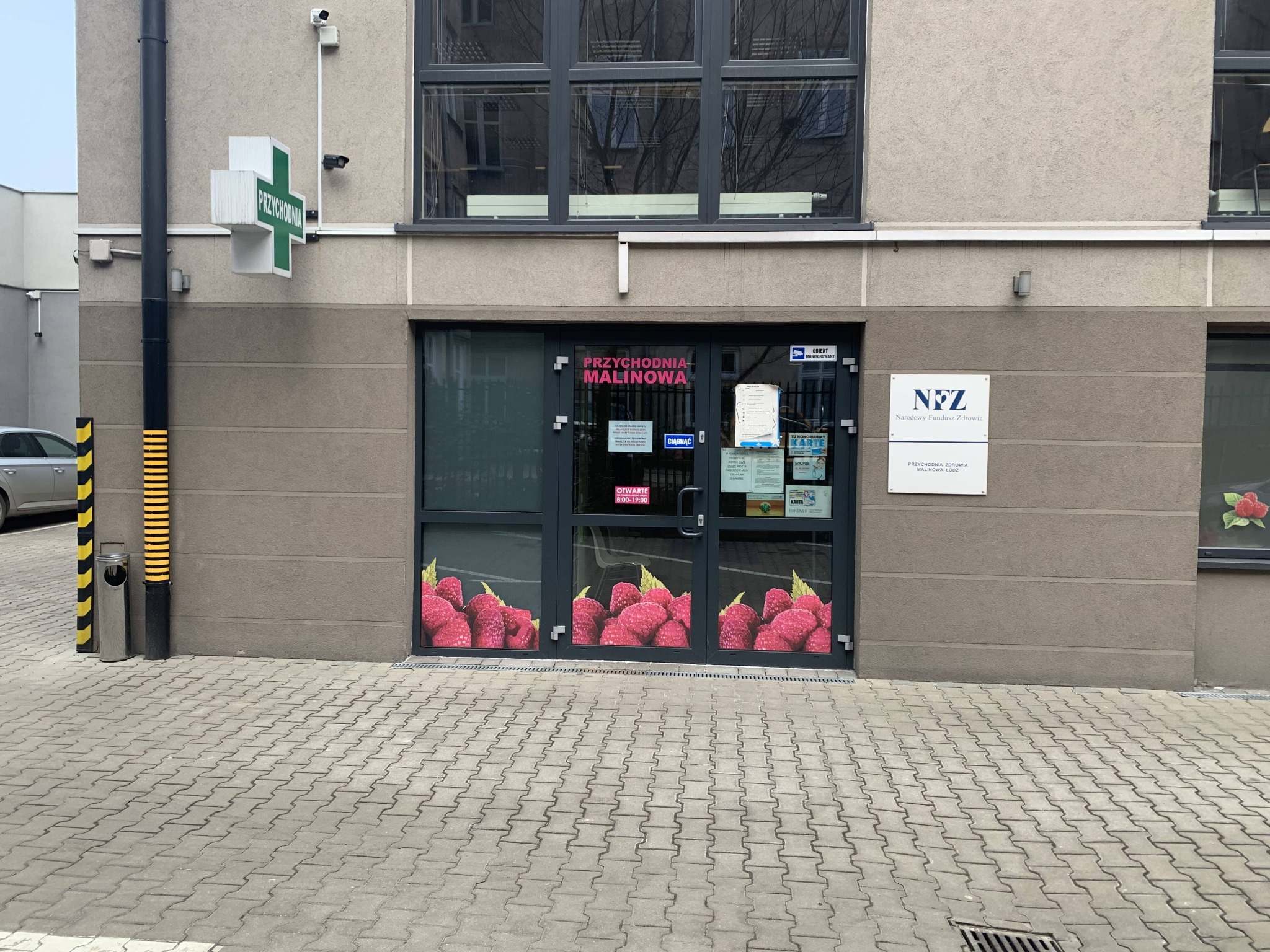 Wejście do przychodni Malinowa z plakatem malin na drzwiach, zielony krzyż apteczny i tabliczka NFZ na fasadzie budynku. Ujęcie z poziomu chodnika.