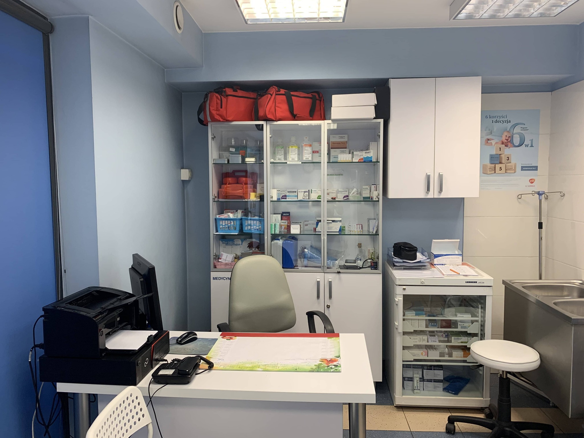 Jasne wnętrze gabinetu lekarskiego z biurkiem, krzesłem, szafkami z lekami i sprzętem medycznym. Widoczne torby medyczne i plakat informacyjny.