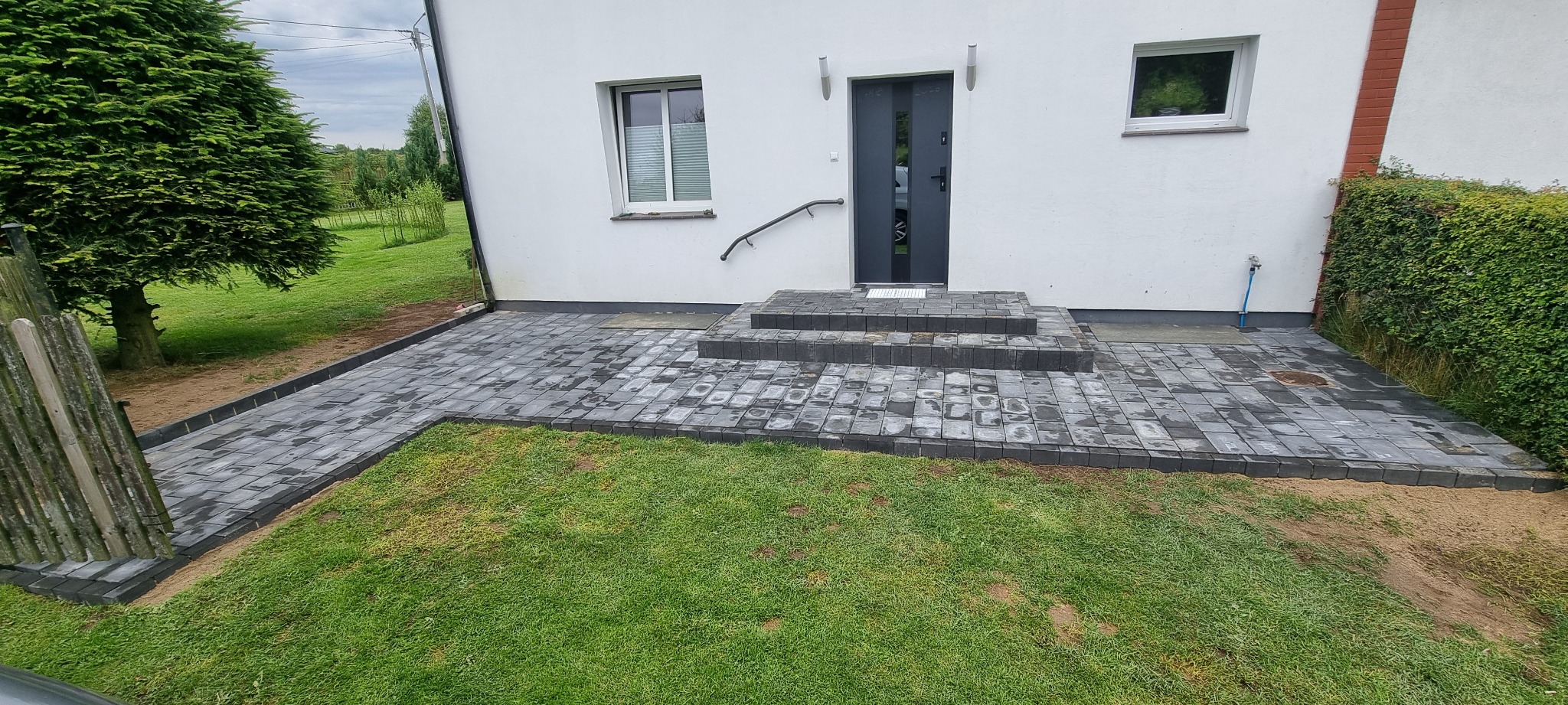 Ułożona kostka brukowa prowadząca do wejścia domu z trzema schodami. Kostka w odcieniach szarości, obrzeża z ciemniejszej kostki. Trawa po bokach.