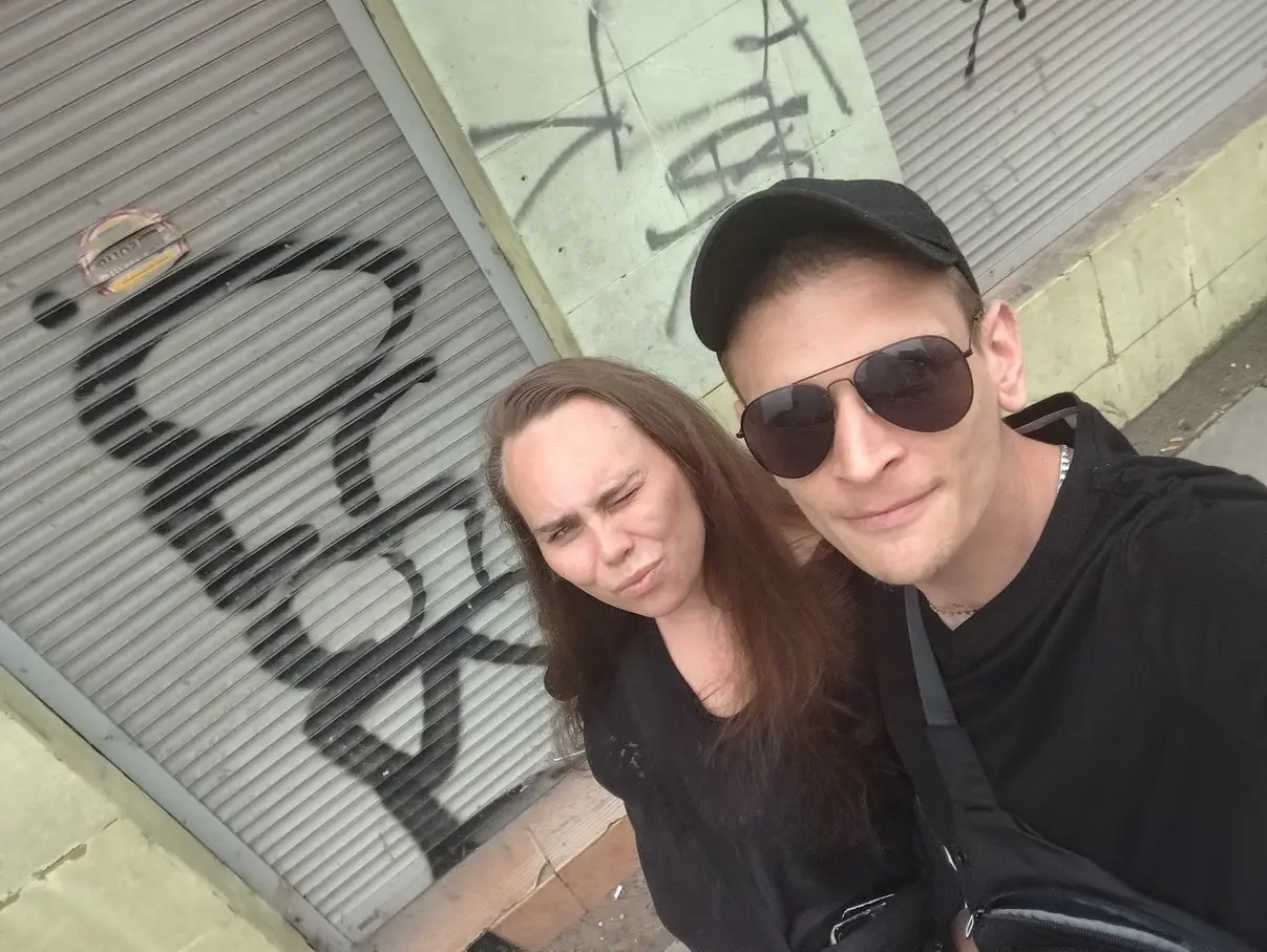 Selfie dwóch osób na tle pomalowanej graffiti, metalowej rolety. Mężczyzna w czapce i okularach, kobieta z długimi włosami robi dziwną minę. Ujęcie z dołu.