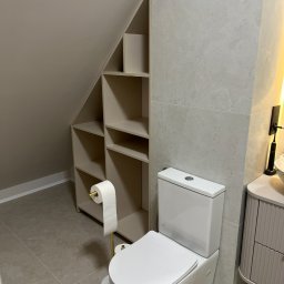 Richmont meble - Jasna łazienka z toaletą i szafką wnękową pod skosem. Beżowe płytki i ściany. Stojak na papier toaletowy w kolorze złotym. Minimalistyczny design.