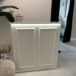 Richmont meble - Biała lada recepcyjna z oświetleniem LED, dekorowana ramkami, z kwiatami i elementami wystroju w tle. Minimalistyczny design, jasne wnętrze.