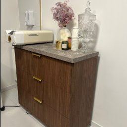 Richmont meble - Komoda z trzema szufladami w odcieniach brązu, z blatem imitującym kamień. Na blacie dekoracje: kwiaty, świece, szklany dystrybutor wody i kubeczki. Minimalistyczny design.