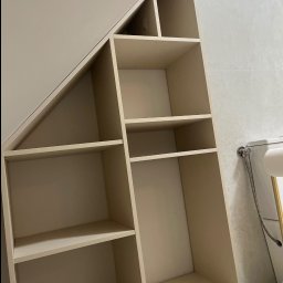 Richmont meble - Beżowa, zabudowana szafa wnękowa z półkami, dopasowana do skosu dachu, widoczna w łazience z jasnymi płytkami. Minimalistyczny design, funkcjonalne wykorzystanie przestrzeni.