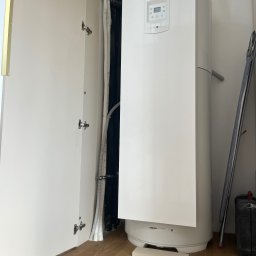 Richmont meble - Biały bojler Atlantic w szafie z otwartymi drzwiami, widoczne rury i elementy instalacji. Podłoga z paneli, drabina oparta o ścianę, skrzynka narzędziowa obok.