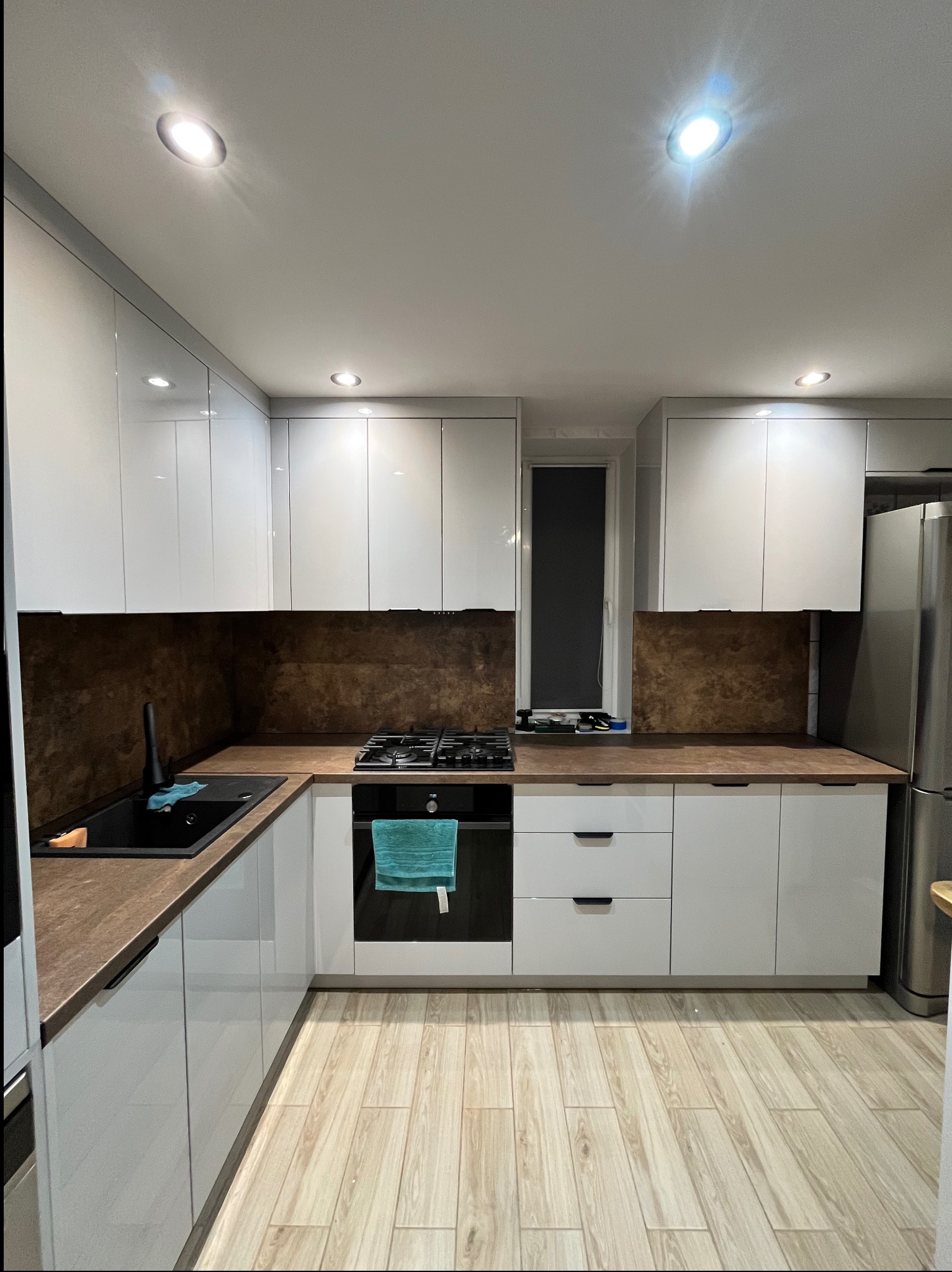 Nowoczesna kuchnia na wymiar z białymi, lakierowanymi frontami, brązowym blatem i płytkami imitującymi rdzę. Uwagę zwraca czarny zlew i bateria.