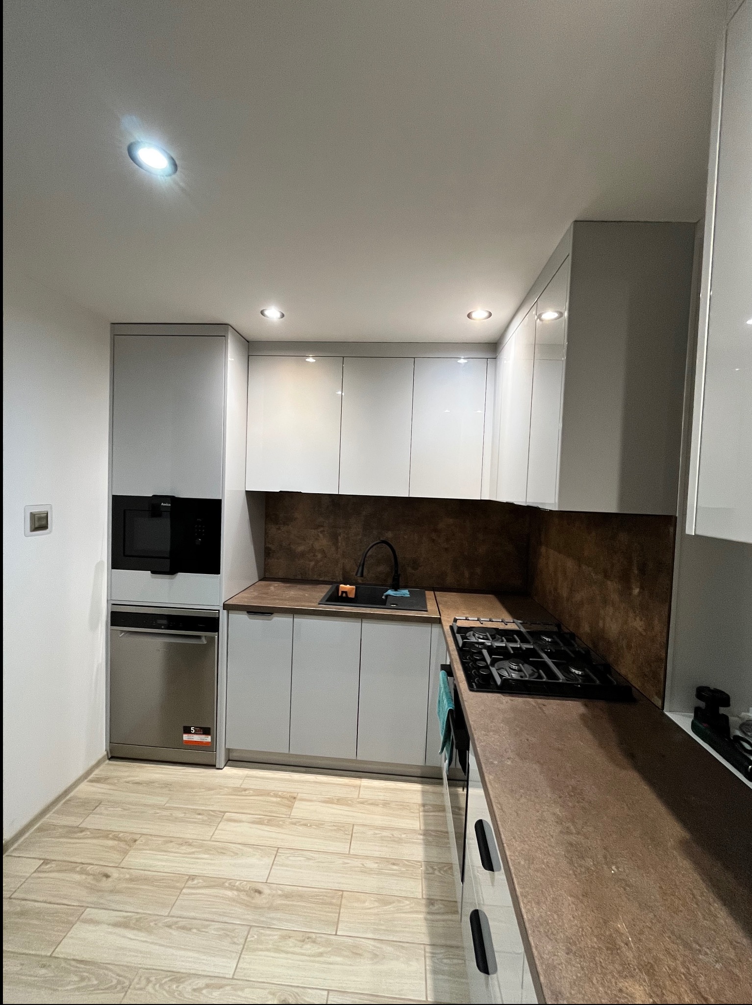 Jasna kuchnia z drewnianym blatem i sprzętami AGD w zabudowie. Uwagę zwraca ciemnobrązowy panel nad blatem i płyta gazowa.