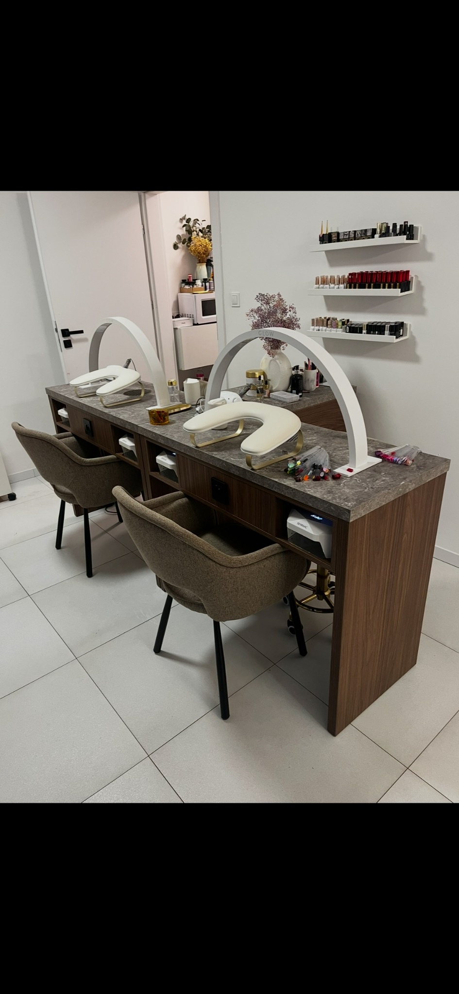 Dwa stanowiska do manicure z lampami LED, krzesłami i półkami na lakiery. Widoczne akcesoria do stylizacji paznokci i dekoracje. Jasne wnętrze salonu kosmetycznego.