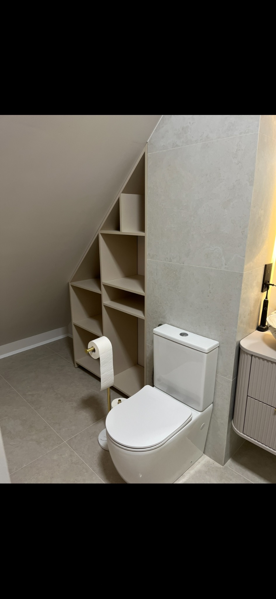 Jasna łazienka z toaletą i szafką wnękową pod skosem. Beżowe płytki i ściany. Stojak na papier toaletowy w kolorze złotym. Minimalistyczny design.