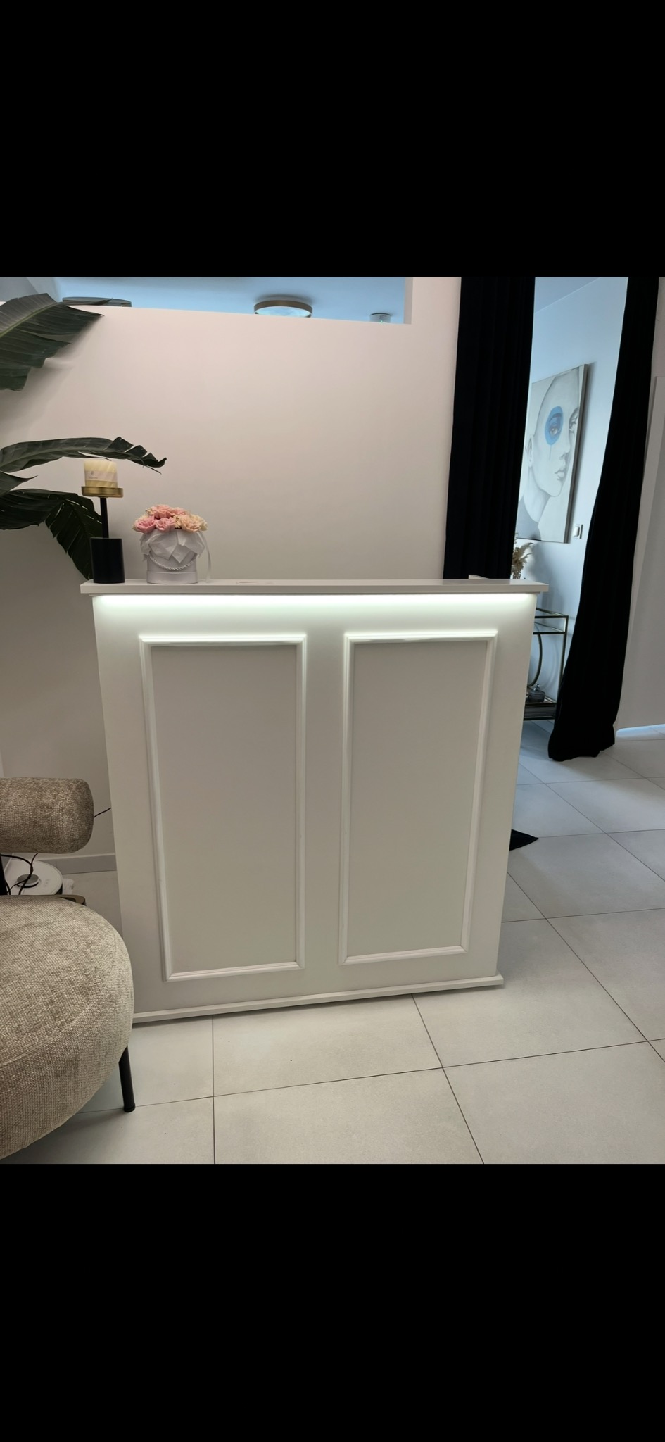 Biała lada recepcyjna z oświetleniem LED, dekorowana ramkami, z kwiatami i elementami wystroju w tle. Minimalistyczny design, jasne wnętrze.