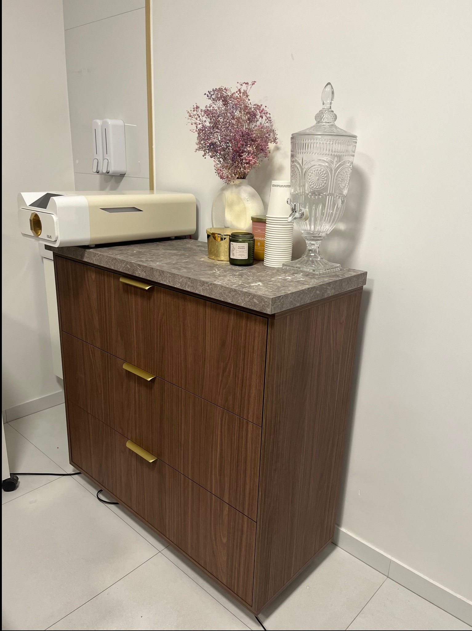 Komoda z trzema szufladami w odcieniach brązu, z blatem imitującym kamień. Na blacie dekoracje: kwiaty, świece, szklany dystrybutor wody i kubeczki. Minimalistyczny design.