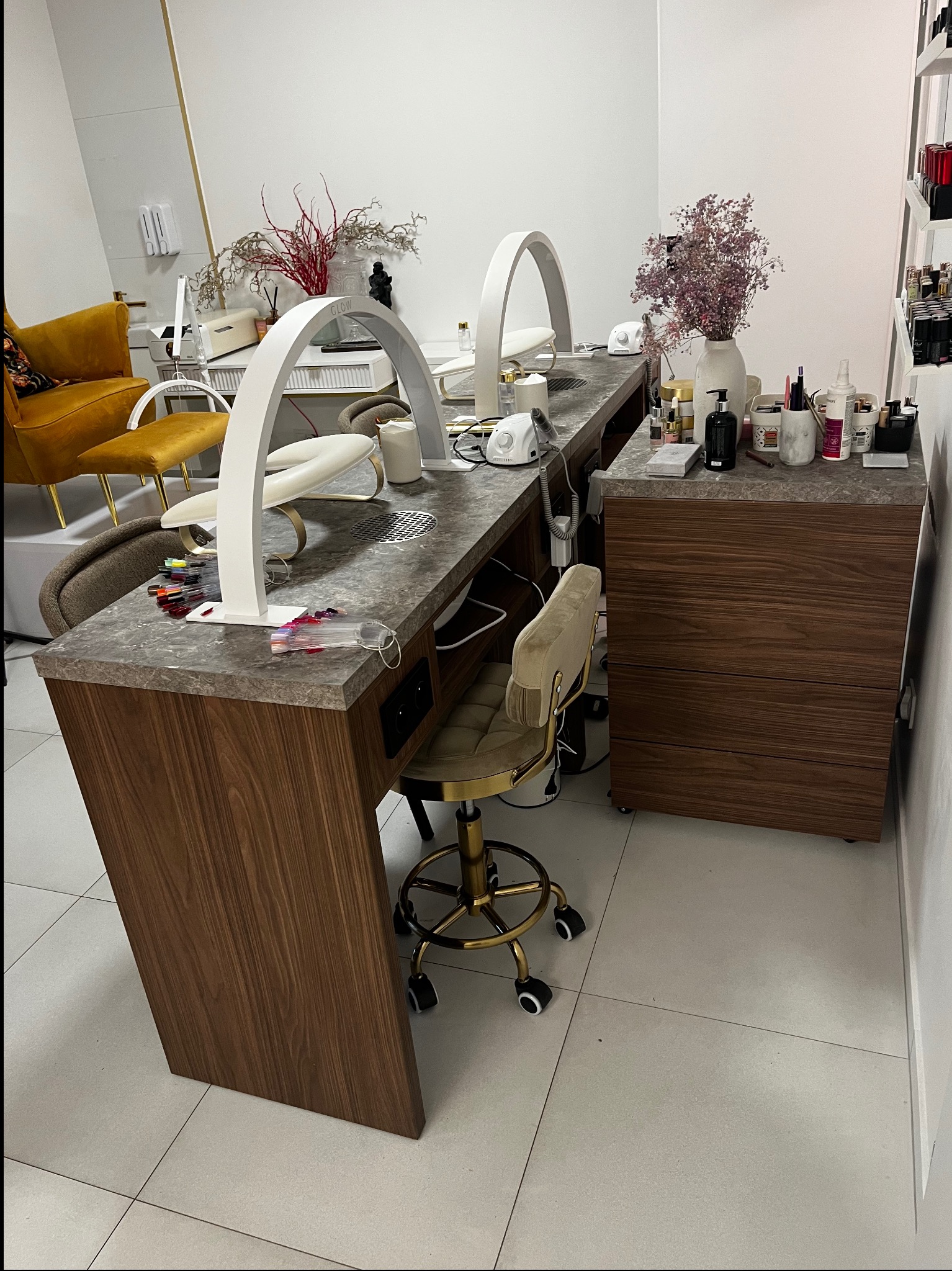 Długi, drewniany stół do manicure z marmurowym blatem, oświetleniem LED w kształcie łuku i krzesłami na złotych kółkach. Widoczne akcesoria do manicure i dekoracje.