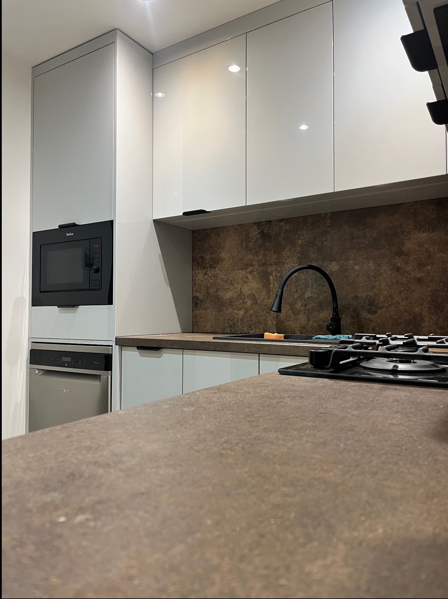 Nowoczesna kuchnia z szarymi szafkami, brązowym blatem i sprzętem AGD w zabudowie. Ujęcie z perspektywy blatu, z widoczną płytą grzewczą i baterią.