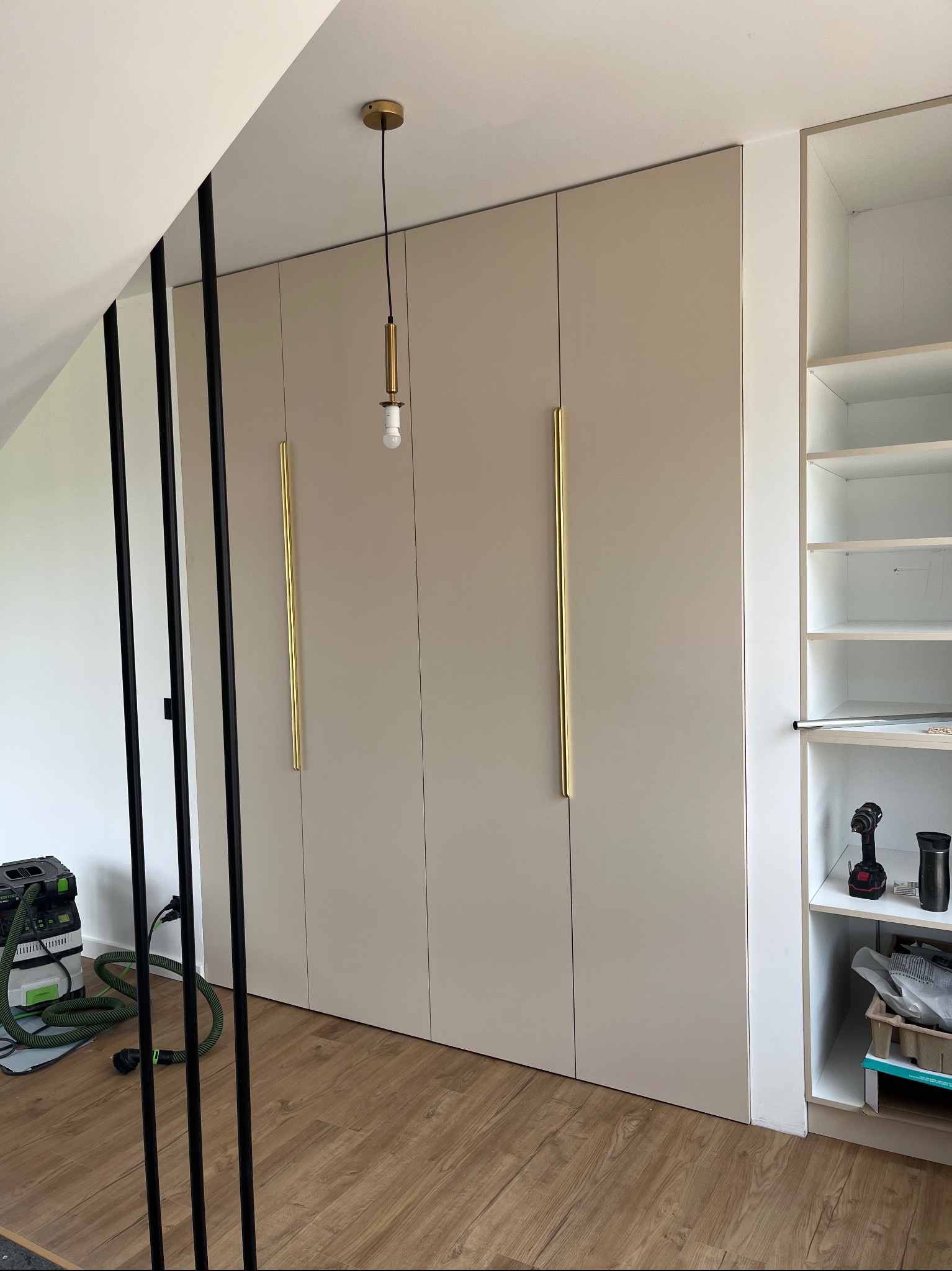 Beżowa szafa na wymiar z długimi, złotymi uchwytami, oświetleniem i otwartą półką z narzędziami, na drewnianej podłodze. Widoczny odkurzacz przemysłowy.