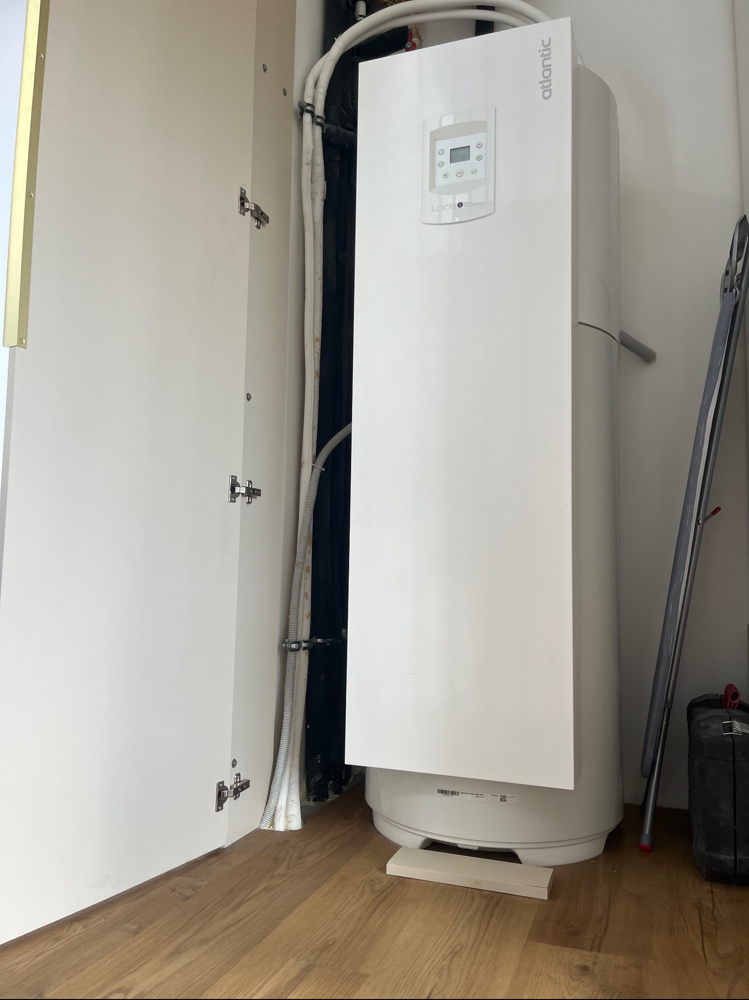 Biały bojler Atlantic w szafie z otwartymi drzwiami, widoczne rury i elementy instalacji. Podłoga z paneli, drabina oparta o ścianę, skrzynka narzędziowa obok.