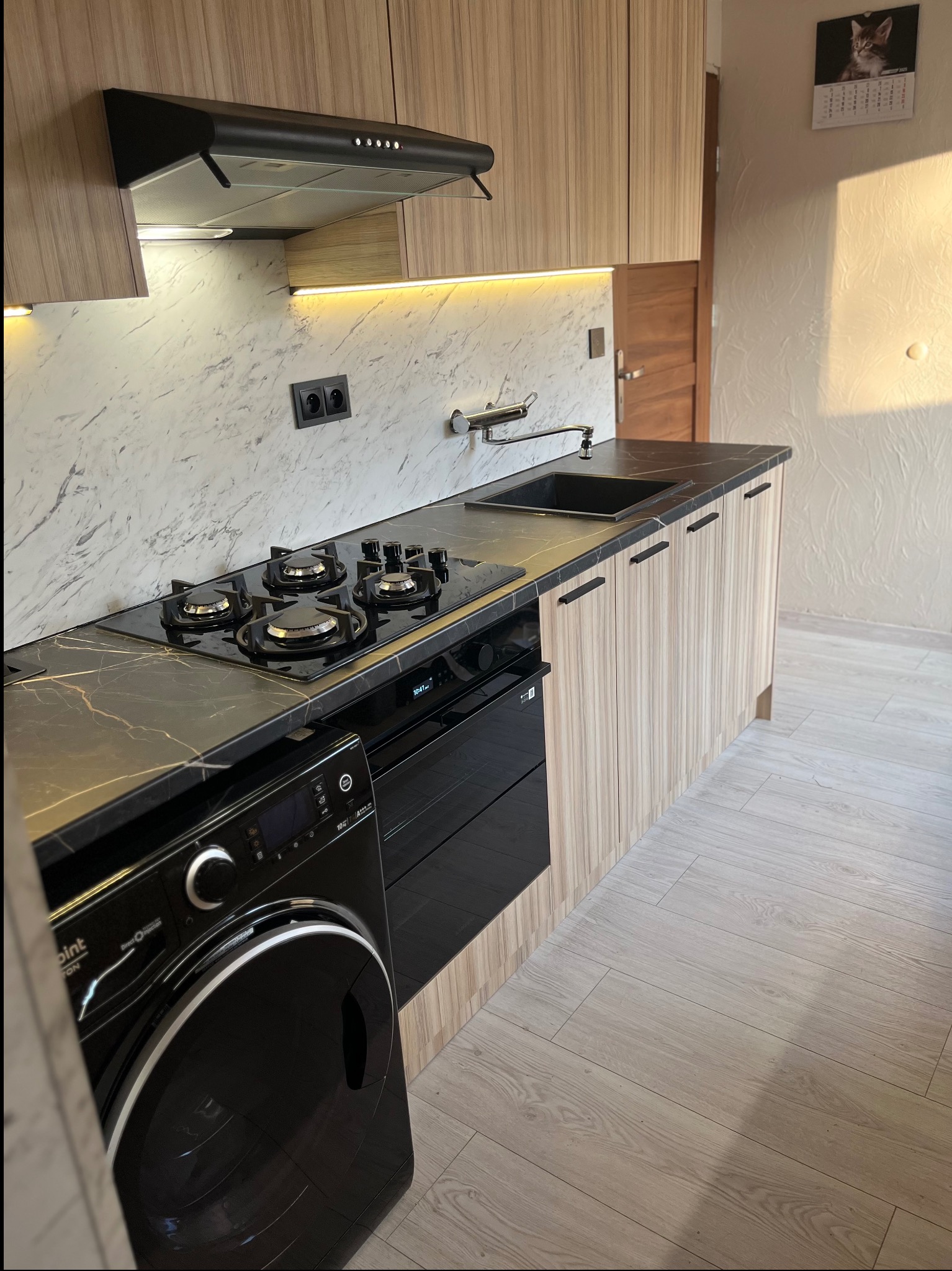 Nowoczesna kuchnia z blatem imitującym marmur, czarną płytą grzewczą i piekarnikiem. Jasne, drewnopodobne fronty szafek i czarny okap. Na ścianie kalendarz z kotem.