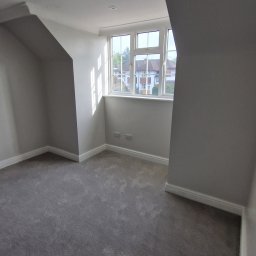 DRW - Dokładne Remonty Wykonczenia - Jasne, świeżo pomalowane poddasze z szarym dywanem i oknem z widokiem na domy. Ściany w odcieniach szarości, listwy przypodłogowe i sufit w bieli. Minimalistyczny wystrój.