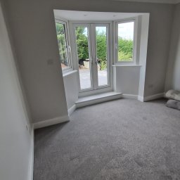 DRW - Dokładne Remonty Wykonczenia - Jasne wnętrze pokoju po remoncie z szarą wykładziną i białymi ścianami. Widoczne okno balkonowe z widokiem na zieleń. Minimalistyczny design, świeżo po malowaniu.