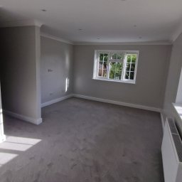 DRW - Dokładne Remonty Wykonczenia - Jasne, świeżo pomalowane wnętrze pokoju z szarymi ścianami, białymi listwami przypodłogowymi i oknami. Widoczny grzejnik pod oknem i miękka wykładzina podłogowa.