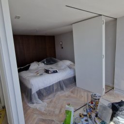 DRW - Dokładne Remonty Wykonczenia - Pokój w trakcie malowania: zabezpieczone łóżko folią, widoczne narzędzia malarskie, świeżo pomalowane ściany na biało, drewniana podłoga w jodełkę.