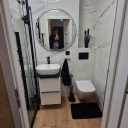 DRW - Dokładne Remonty Wykonczenia - Nowoczesna łazienka z kabiną prysznicową typu walk-in, umywalką na szafce, okrągłym lustrem i drewnopodobną podłogą. Marmurowe płytki na ścianach.