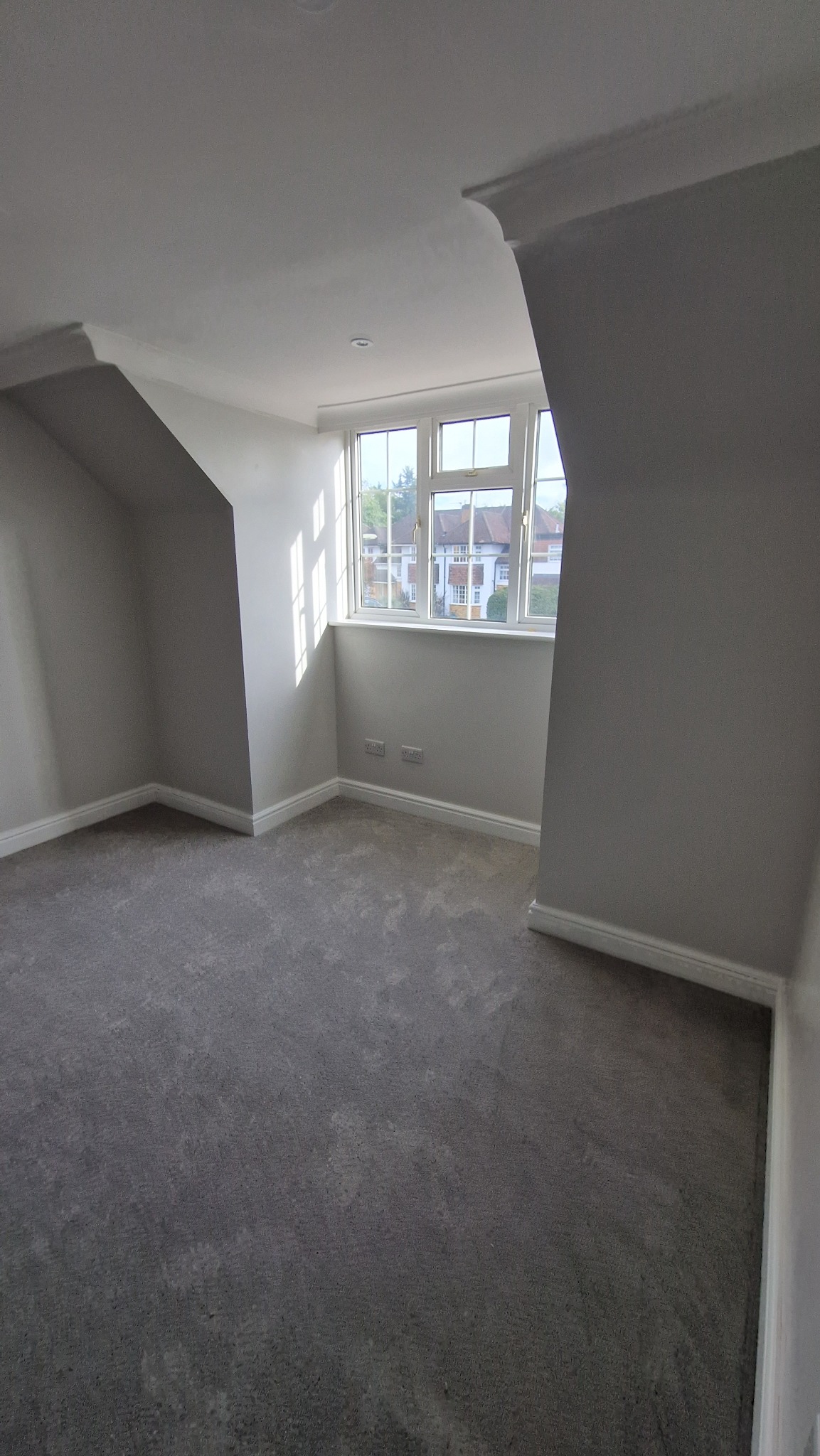 Jasne, świeżo pomalowane poddasze z szarym dywanem i oknem z widokiem na domy. Ściany w odcieniach szarości, listwy przypodłogowe i sufit w bieli. Minimalistyczny wystrój.