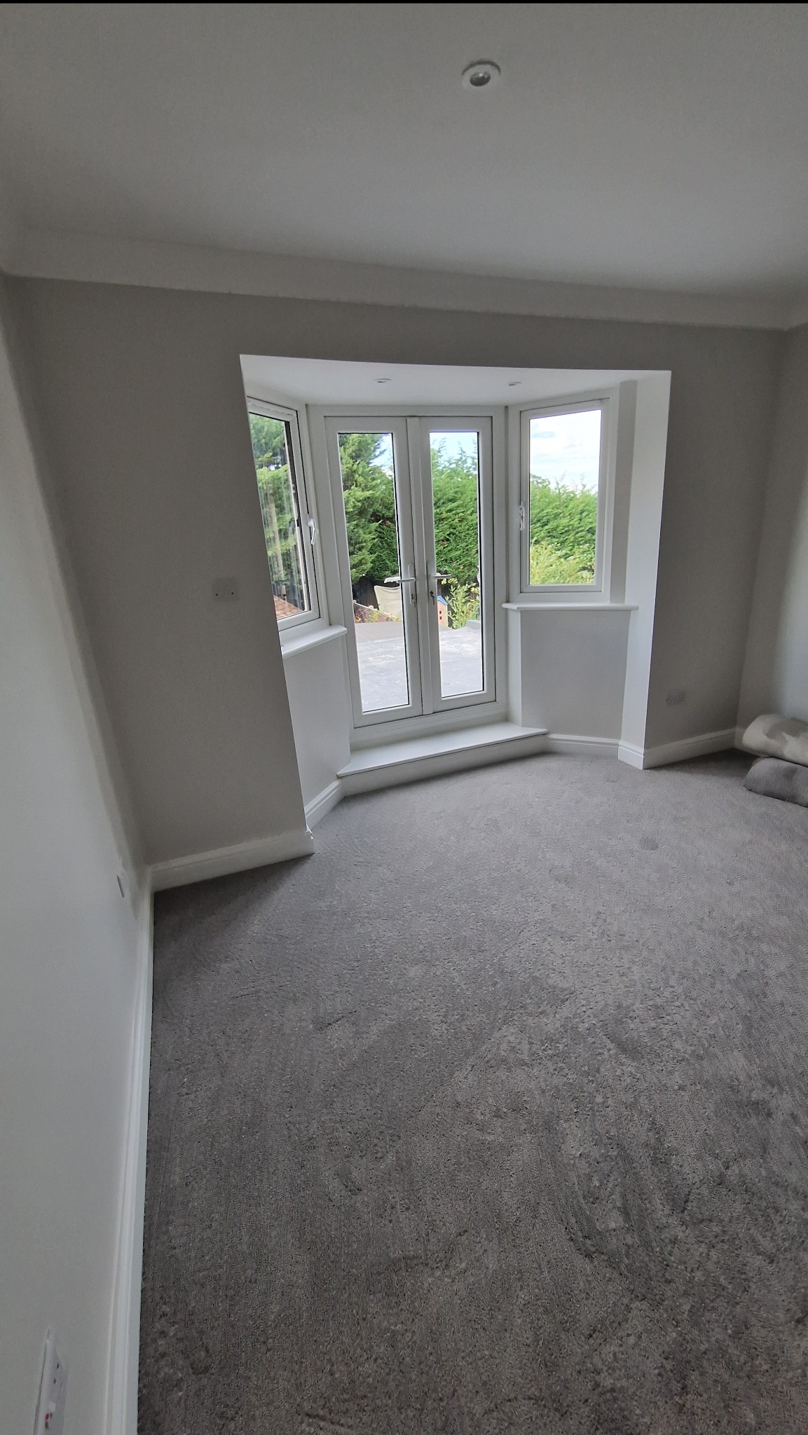 Jasne wnętrze pokoju po remoncie z szarą wykładziną i białymi ścianami. Widoczne okno balkonowe z widokiem na zieleń. Minimalistyczny design, świeżo po malowaniu.