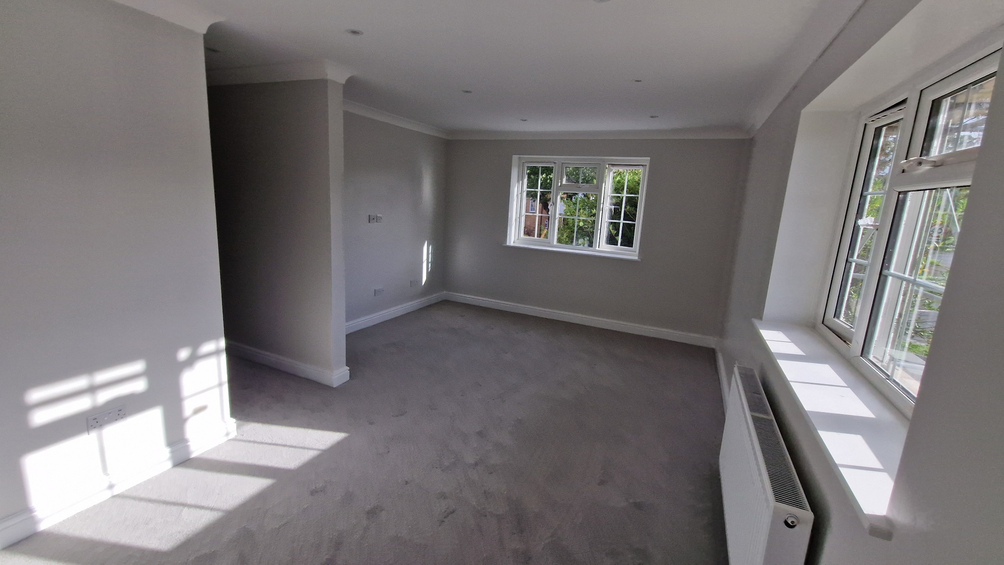 Jasne, świeżo pomalowane wnętrze pokoju z szarymi ścianami, białymi listwami przypodłogowymi i oknami. Widoczny grzejnik pod oknem i miękka wykładzina podłogowa.