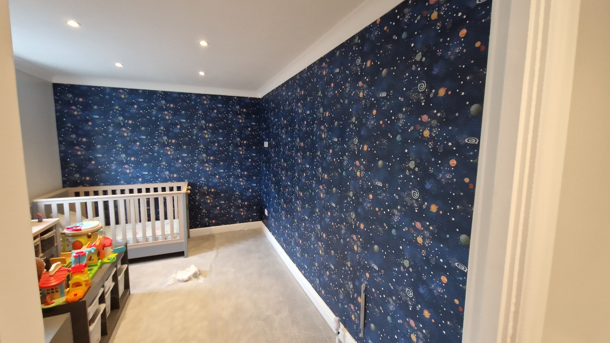 Pokój dziecięcy po remoncie: ściany pokryte tapetą w kosmiczne wzory, biała listwa przysufitowa, łóżeczko i zabawki. Nowoczesne oświetlenie sufitowe.