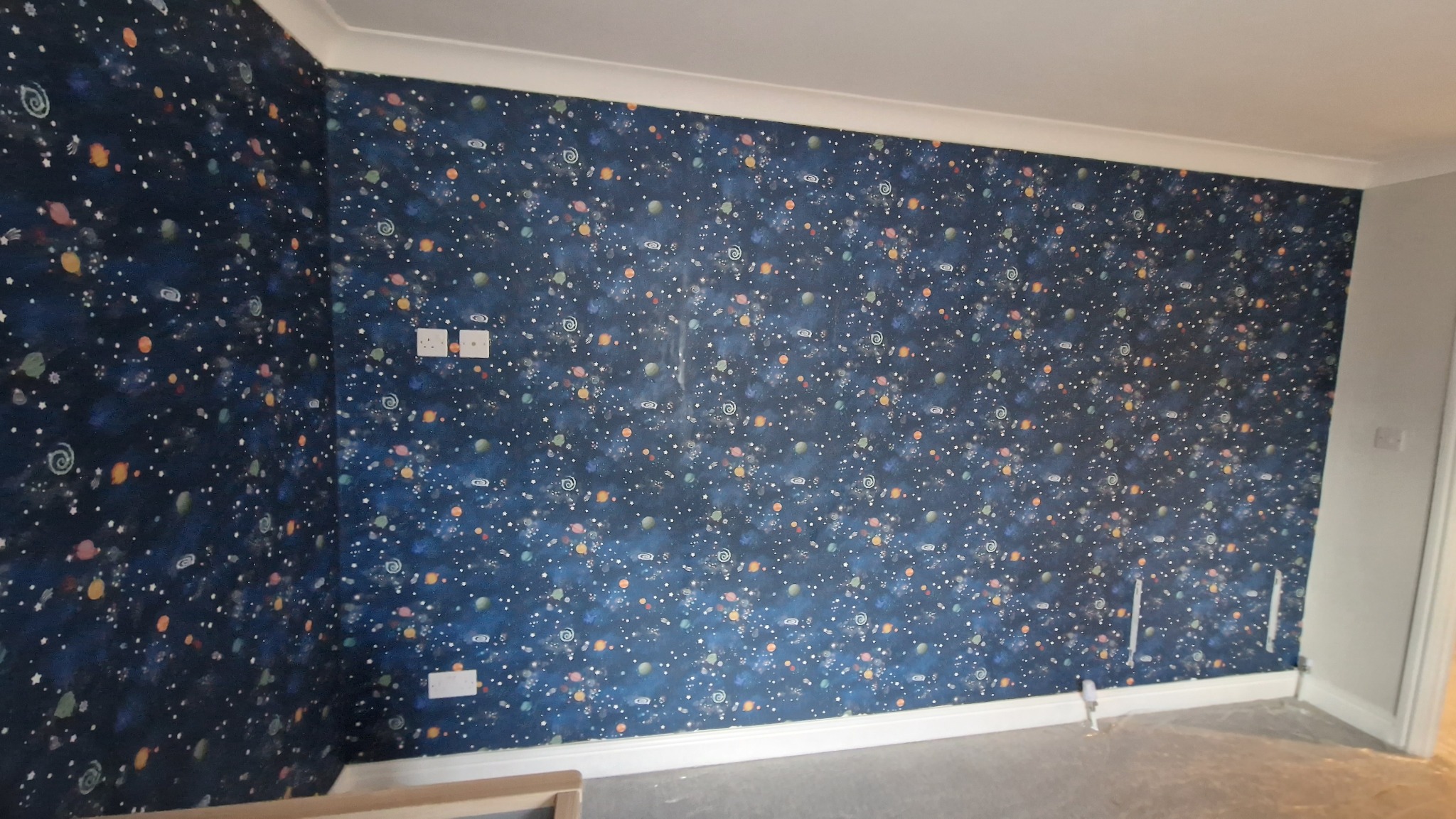 Ściana w pokoju dziecięcym po malowaniu, pokryta tapetą w motywy kosmiczne: planety, gwiazdy i spirale na ciemnoniebieskim tle. Widoczne gniazdka elektryczne i listwa przypodłogowa.